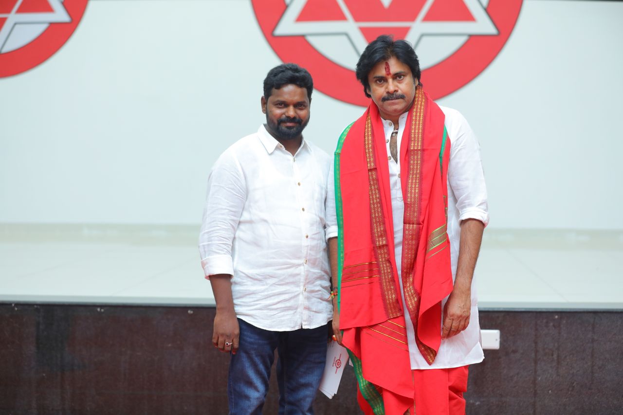 Two Indian men indoors, one in traditional attire, with a logo background.
